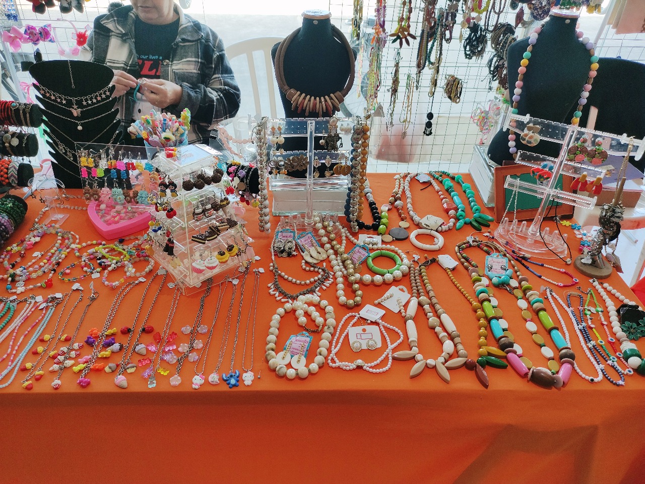Close de uma seleção de colares, pulseiras e brincos sobre uma toalha
laranja, com diversos materiais como pérolas, miçangas coloridas e pingentes temáticos
(cupcakes, ursinhos), e uma artesã ajustando uma peça ao fundo