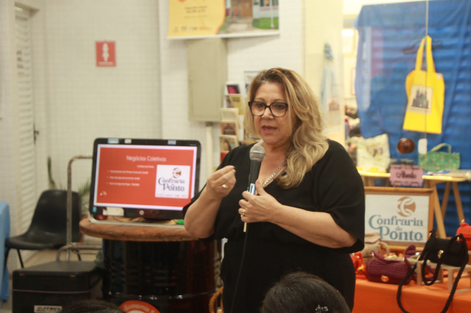 Mulher com cabelos loiros e ondulados, usa óculos de grau e está segurando um microfone enquanto fala para um grupo de pessoas em um ambiente interno. Ela veste uma blusa preta e demonstra expressão séria e envolvida. Ao fundo, uma televisão exibe uma apresentação com o título “Negócios Coletivos” e o logotipo da 