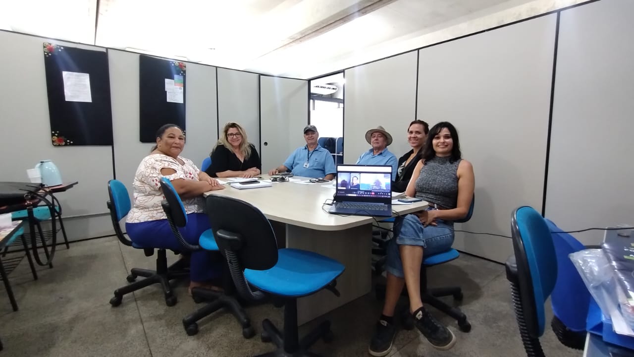 A imagem mostra seis pessoas sentadas ao redor de uma mesa oval em uma sala de reuniões. O grupo, composto por produtores, representantes institucionais e apoiadores do evento, participa de um momento de organização e articulação para a Feira Regional de Produtores Agroecológicos. Sobre a mesa, documentos, cadernos, canetas e um notebook com participantes em videoconferência reforçam o ambiente de trabalho conjunto. O clima é descontraído, com sorrisos que evidenciam o engajamento coletivo e a força da articulação territorial