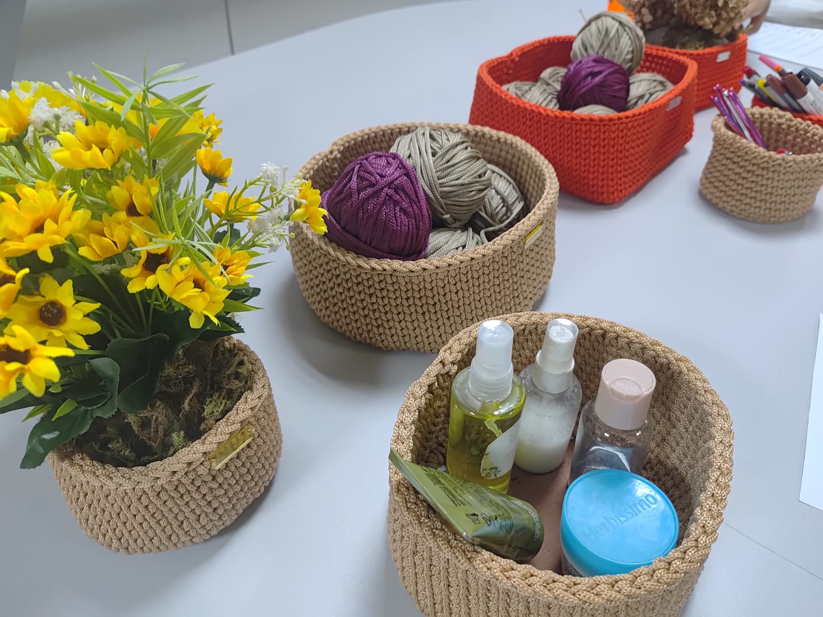 Vista aproximada de quatro cachepôs de crochê em diferentes tamanhos e cores – bege, laranja e cru – dispostos sobre uma mesa branca. Um dos cestos contém flores artificiais amarelas, outro guarda novelos de fios e um terceiro exibe potes de cosméticos artesanais.
